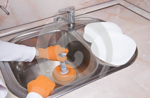 WomanÃ¢â¬â¢s hands with orange gloves cleaning sewer at kitchen faucet over metal sink. hand with plunger